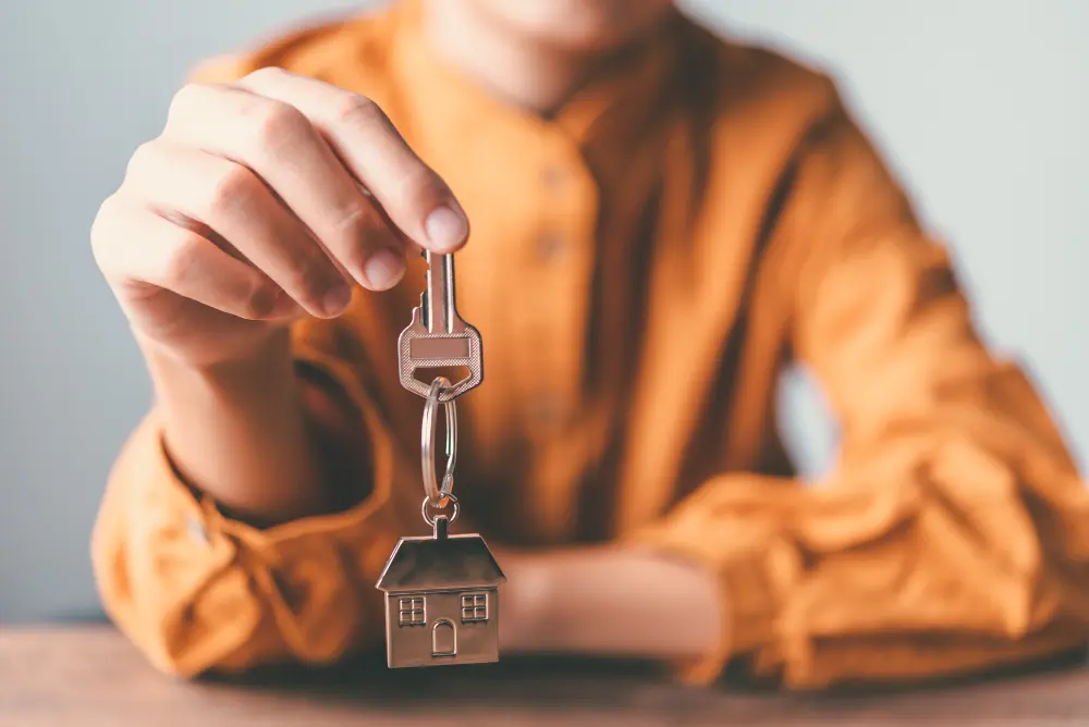 Person in an orange shirt holds a key with a small house-shaped pendant, symbolizing home ownership or a new home.