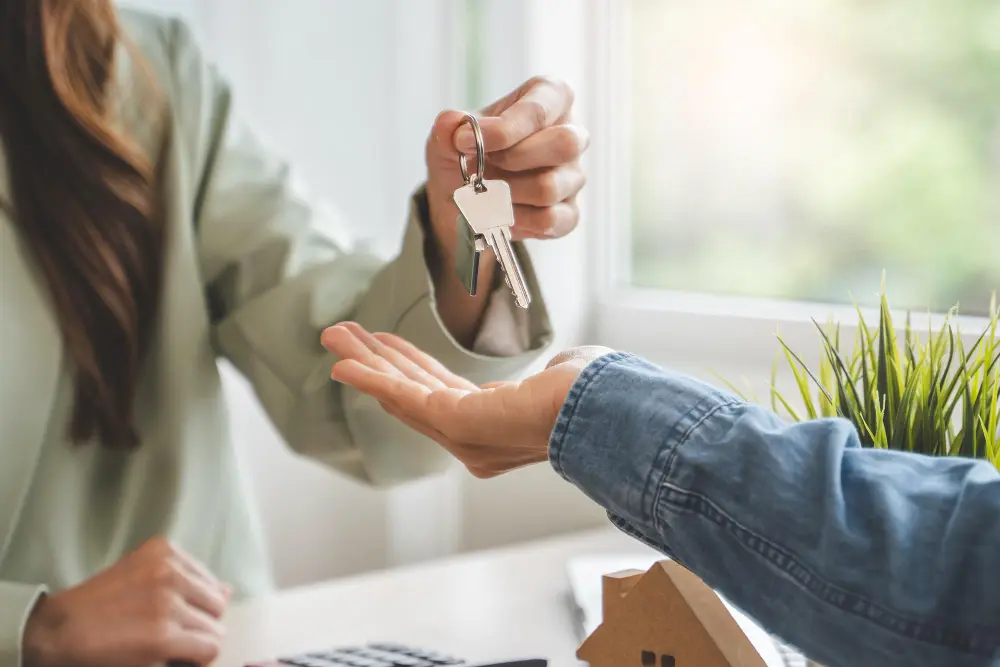 alarma habitatge nou - Protegim - Protegim Person hands over a set of house keys to another person at a desk, suggesting a home sale or rental.